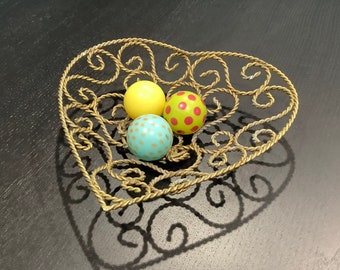 May include: A heart-shaped, gold-colored wire basket holds three decorative spheres. One sphere is yellow, another is green with red polka dots, and the third is turquoise with white dots. The basket sits on a dark surface.