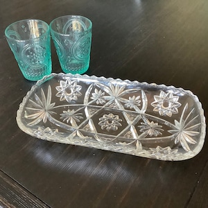 May include: Two turquoise drinking glasses with embossed patterns sit next to a clear, rectangular glass serving dish. The dish features a starburst and floral design. The items are arranged on a dark wooden surface.