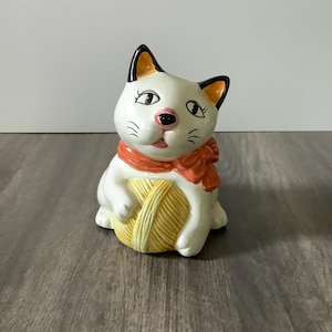 May include: A ceramic cat figurine, white with black and orange accents, wearing an orange scarf and holding a yellow yarn ball. The cat has detailed facial features, including a pink nose and black whiskers. The figurine is on a wooden surface.