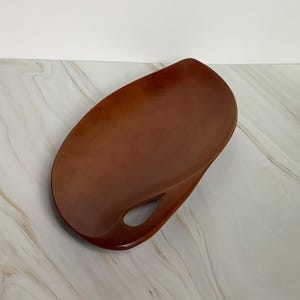 May include: A brown, teardrop-shaped wooden tray with a handle cutout. The tray has a smooth, polished surface and a warm, natural wood tone. The tray is empty and sits on a marble-patterned surface.
