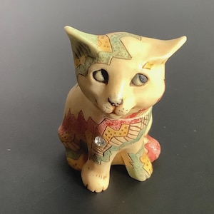 May include: A ceramic cat figurine with a cream-colored body and colorful geometric patterns in green, yellow, and red. The cat has a small rhinestone on its chest and is sitting upright with its ears perked.