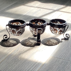 May include: A decorative candle holder with three glass bowls. The bowls are brown and speckled, held by a dark metal frame with curved accents. The candle holder sits on a light-colored surface.