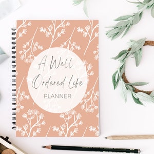 May include: A pink planner with a white floral pattern and the text "A Well Ordered Life Planner" in a white circle.