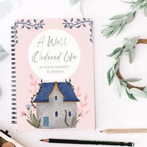May include: A pink spiral bound planner with a white circle on the cover that says "A Well Ordered Life" and "A Homemaker's Planner". The cover also features a watercolor illustration of a gray house with a blue roof and a blue door.