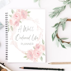 May include: A white planner with a floral design on the cover. The planner has a spiral binding and says "A Well Ordered Life Planner" on the front.