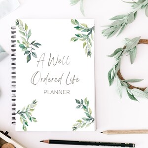 May include: A white spiral bound planner with green watercolor leaves on the cover. The text on the cover reads "A Well Ordered Life Planner".
