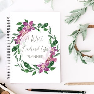May include: A spiral-bound planner with the text "A Well Ordered Life PLANNER" surrounded by a floral wreath of purple flowers and green leaves. A light blue ribbon, a wooden ring with greenery, and pencils are also present.