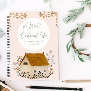 May include: A pink and white spiral bound notebook with the title "A Well Ordered Life: A Homemaker's Planner" and a watercolor illustration of a house with a brown roof.