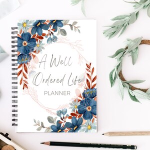 May include: A white planner with a blue and red floral design. The planner has a spiral binding and the text "A Well Ordered Life Planner" on the front cover.