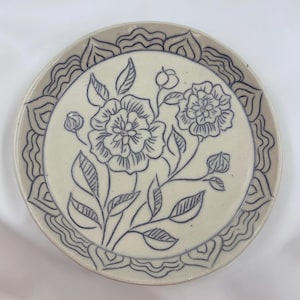 May include: A ceramic plate with a floral design. The plate features a cream-colored center with a blue floral illustration and a decorative border. The flowers and leaves are detailed with fine lines, creating an elegant aesthetic.