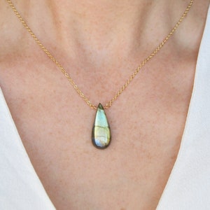 May include: A gold chain necklace with a teardrop-shaped labradorite gemstone pendant. The gemstone has a green, blue, and gold iridescent sheen.