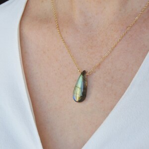 May include: A gold chain necklace with a teardrop-shaped labradorite pendant. The pendant has a dark green, blue, and gold iridescent sheen.