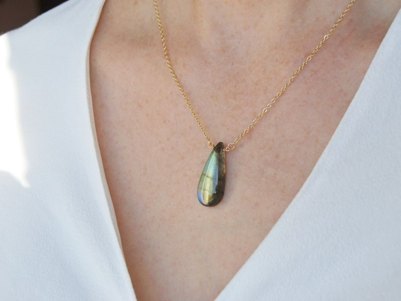 May include: A gold chain necklace with a teardrop-shaped labradorite pendant. The pendant has a dark green, blue, and gold iridescent sheen.
