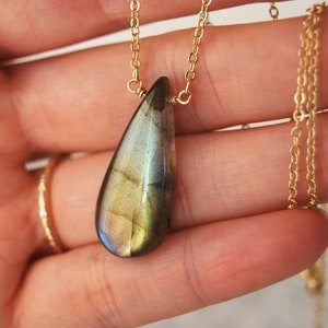 May include: A gold chain necklace with a teardrop-shaped labradorite pendant. The pendant has a green, blue, and gold iridescent sheen.