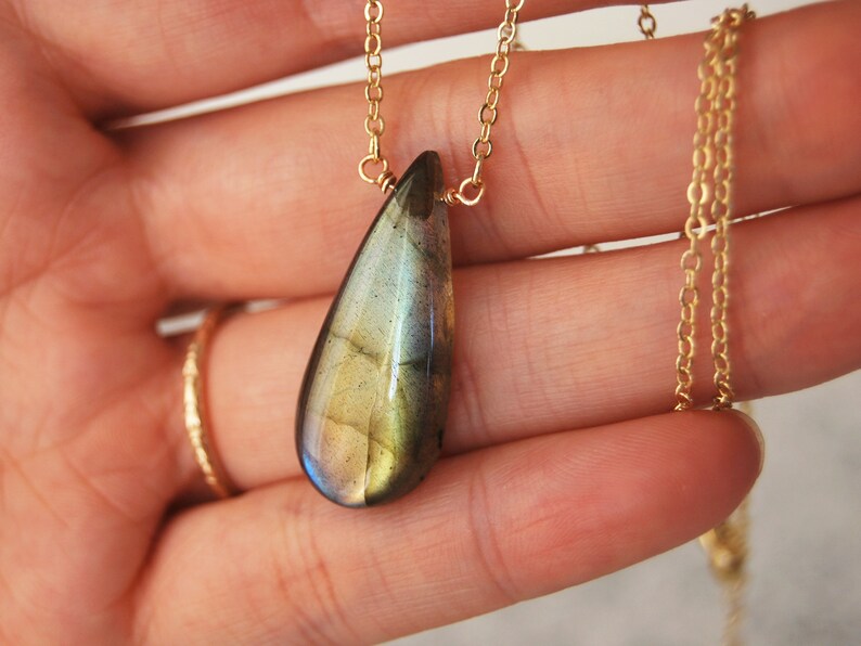 May include: A gold chain necklace with a teardrop-shaped labradorite pendant. The pendant has a green, blue, and gold iridescent sheen.
