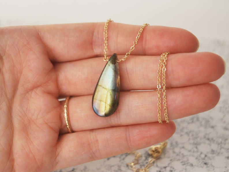 May include: A gold chain necklace with a teardrop-shaped labradorite pendant. The pendant has a green, gold, and black iridescent sheen.