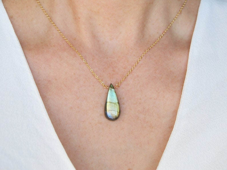 May include: A gold chain necklace with a teardrop-shaped labradorite gemstone pendant. The gemstone has a green, blue, and gold iridescent sheen.