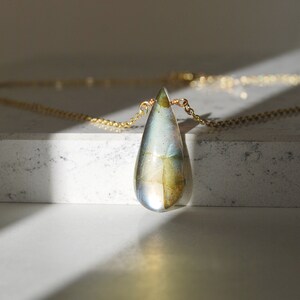 May include: A gold chain necklace with a teardrop-shaped pendant made of iridescent glass. The pendant has a swirling pattern of blue, green, and gold.