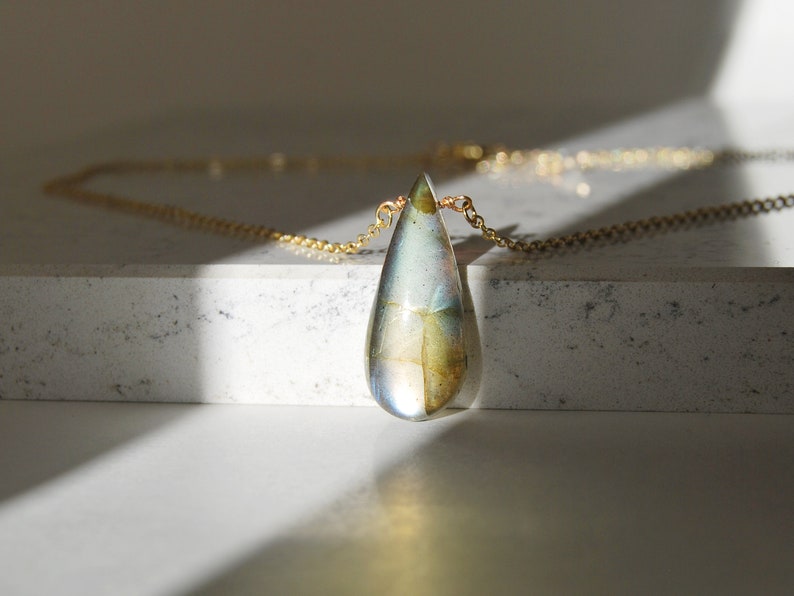 May include: A gold chain necklace with a teardrop-shaped pendant made of iridescent glass. The pendant has a swirling pattern of blue, green, and gold.