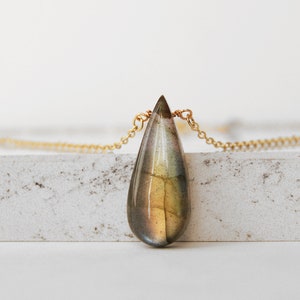 May include: A gold chain necklace with a teardrop-shaped labradorite pendant. The pendant has a green, brown, and gold iridescent sheen.