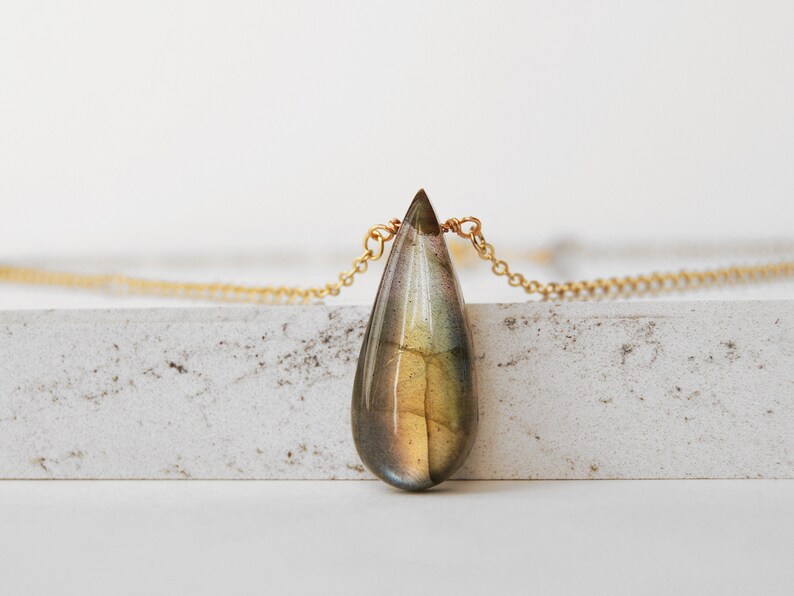 May include: A gold chain necklace with a teardrop-shaped labradorite pendant. The pendant has a green, brown, and gold iridescent sheen.