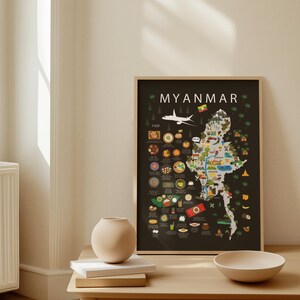 May include: A framed poster showing a map of Myanmar, with illustrations of food, landmarks, and an aeroplane. The word "MYANMAR" is at the top. The poster is on a table with a vase, books, and a bowl.