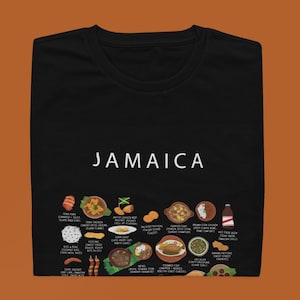 Puede incluir: Camiseta negra con la palabra "JAMAICA" en blanco. Debajo del texto, ilustraciones de varios platos jamaicanos, como pollo jerk, empanadas y ackee y bacalao. La camiseta está doblada.