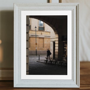 Op de afbeelding: Een ingelijste foto van een persoon die door een smalle, gewelfde doorgang in een stad loopt. De doorgang wordt geflankeerd door gebouwen met gele muren en een lantaarnpaal in het midden. De foto is in een wit frame met een getextureerd oppervlak.