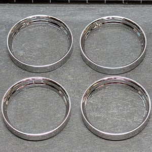 May include: Four silver-colored, circular metal rings are arranged in a square pattern against a dark gray surface. Each ring has a smooth, reflective surface and a layered design, suggesting a decorative or functional purpose.