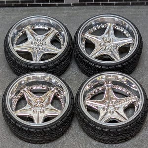 Pode incluir: Quatro rodas de carro cromadas com pneus pretos. Cada roda apresenta um design de estrela de cinco pontas no centro e uma jante polida. Os pneus t&ecirc;m um padr&atilde;o texturizado.