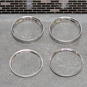 May include: Four silver-colored metal rings are arranged on a gray surface. The rings are circular with a smooth, reflective finish. The background features a dark, textured surface with a mosaic tile pattern.