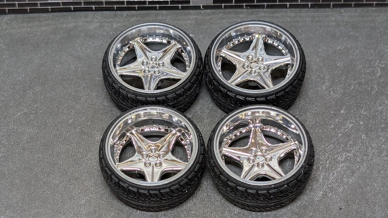 Pode incluir: Quatro rodas de carro cromadas com pneus pretos. Cada roda apresenta um design de estrela de cinco pontas no centro e uma jante polida. Os pneus t&ecirc;m um padr&atilde;o texturizado.
