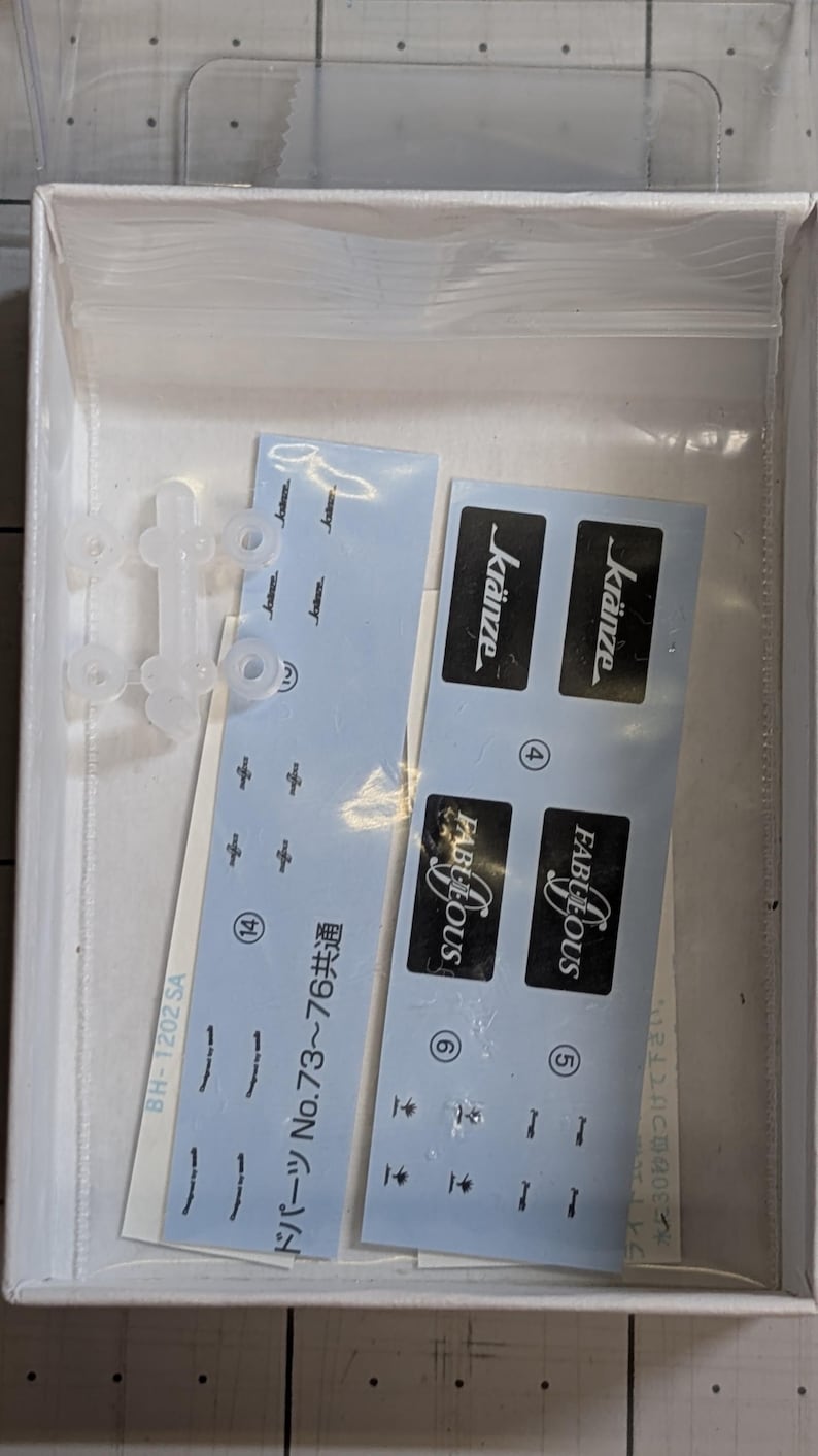 Pode incluir: Uma caixa branca contendo pe&ccedil;as de um kit de modelo. Inclui pe&ccedil;as de pl&aacute;stico transparente, decalques azuis com as palavras "FABULOUS" e "kr&auml;nze" e texto japon&ecirc;s. O kit parece ser para um carro ou ve&iacute;culo modelo.