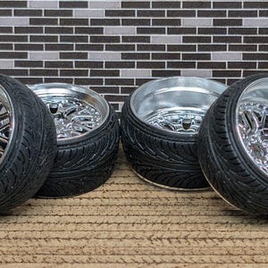 May include: Four black tires with chrome rims are arranged on a brown surface. The rims have a complex, star-like spoke design. The tires have a textured sidewall. The background features a brick-like pattern in shades of gray and black.