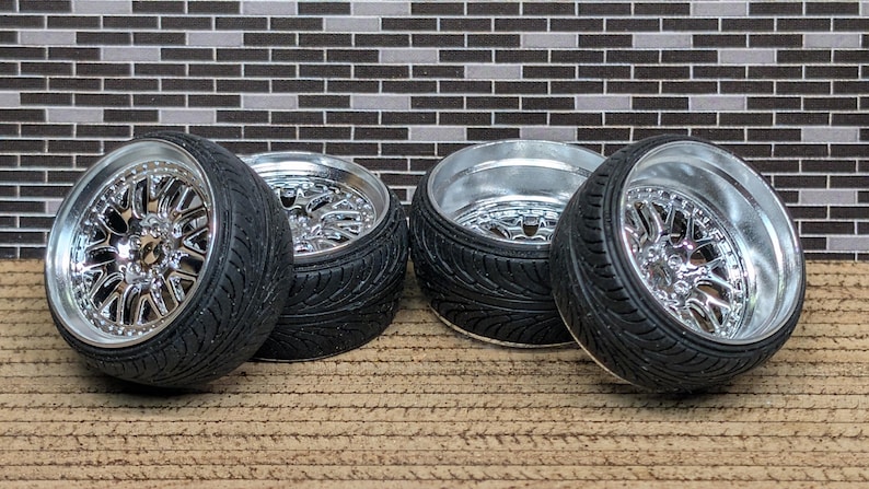 May include: Four black tires with chrome rims are arranged on a brown surface. The rims have a complex, star-like spoke design. The tires have a textured sidewall. The background features a brick-like pattern in shades of gray and black.