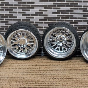 May include: Four black tires with chrome rims. The rims have a multi-spoke design with a polished finish. The tires are arranged in a row against a brick-patterned background.