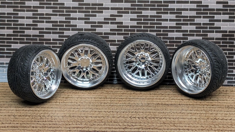 May include: Four black tires with chrome rims. The rims have a multi-spoke design with a polished finish. The tires are arranged in a row against a brick-patterned background.