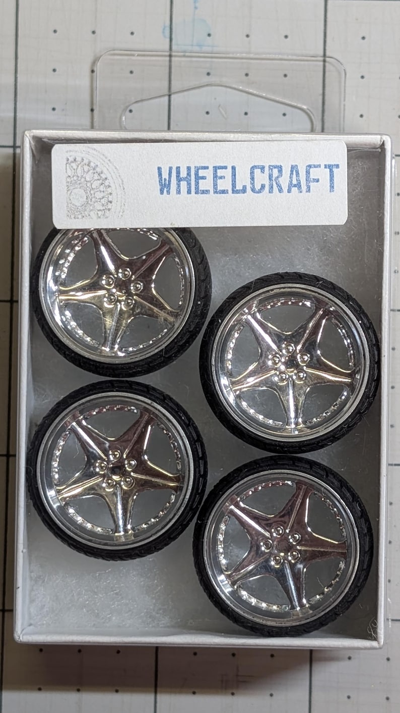 Pode incluir: Quatro rodas de carro em miniatura com acabamento cromado e pneus pretos, apresentadas numa embalagem de pl&aacute;stico transparente. As rodas t&ecirc;m um design de cinco raios e est&atilde;o rotuladas como "WHEELCRAFT" numa etiqueta branca.