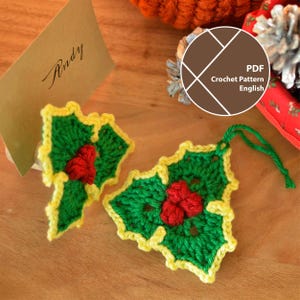 May include: Two handmade crochet holly leaf decorations with red berries and yellow edging. One has a green hanging loop. A brown card with the name "Andy" is in the background. The image also includes a PDF crochet pattern.