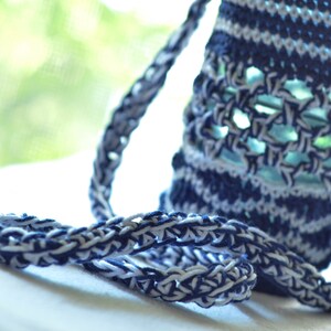 May include: Close-up of a handmade, crocheted bag in shades of navy blue and white. The bag features a textured, open weave design with a braided strap. The background is blurred, with a glimpse of green foliage.