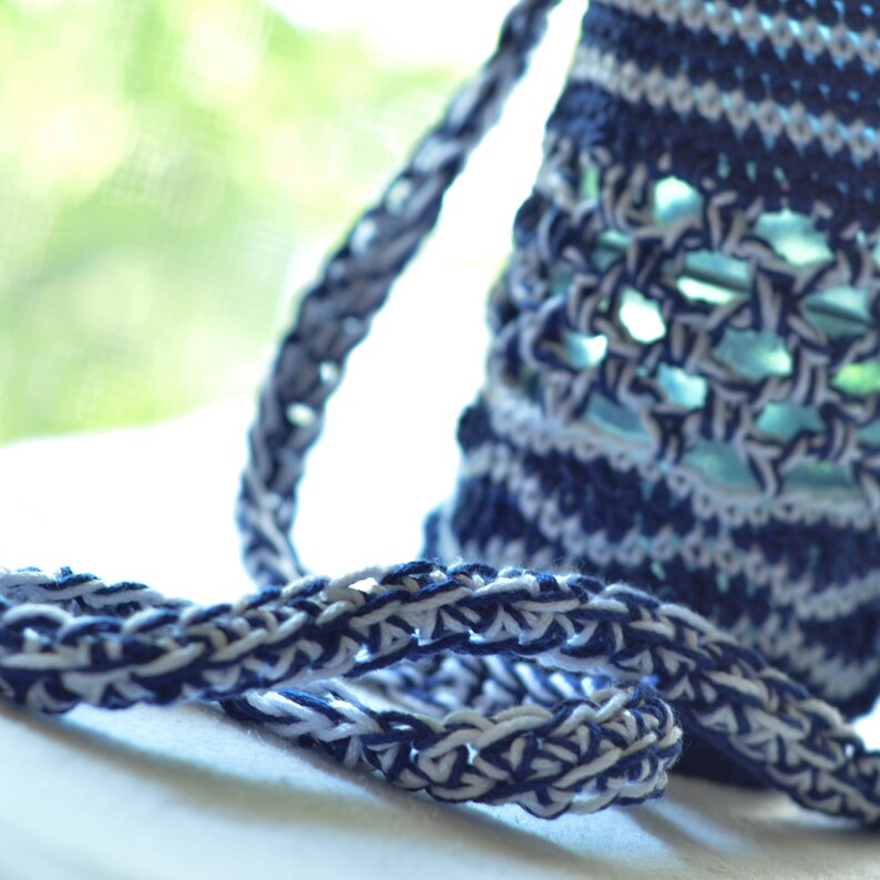 May include: Close-up of a handmade, crocheted bag in shades of navy blue and white. The bag features a textured, open weave design with a braided strap. The background is blurred, with a glimpse of green foliage.