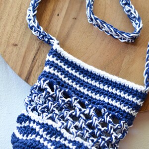 May include: A handmade crochet bag in navy blue and white. The bag features a woven pattern with a matching strap. The bag is sitting on a wooden surface.