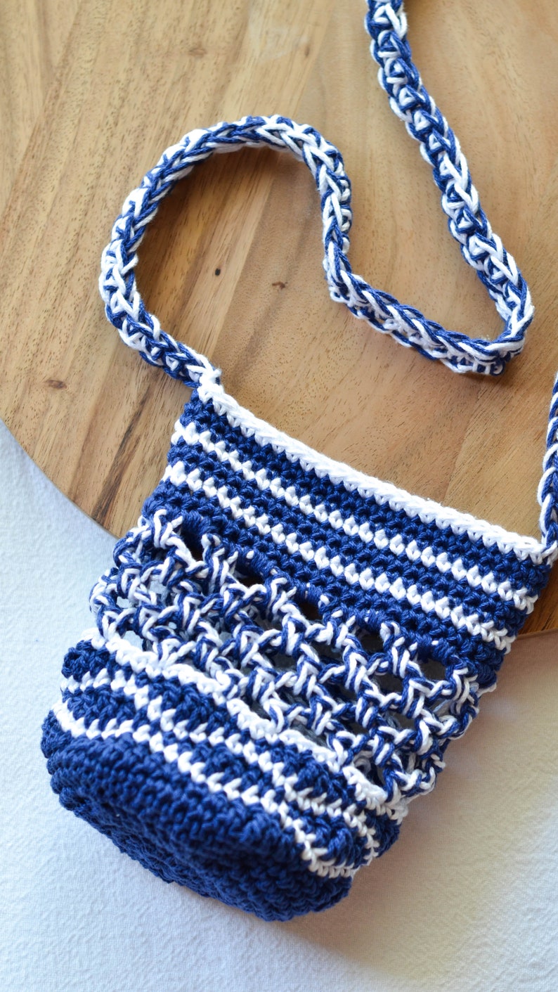 May include: A handmade crochet bag in navy blue and white. The bag features a woven pattern with a matching strap. The bag is sitting on a wooden surface.