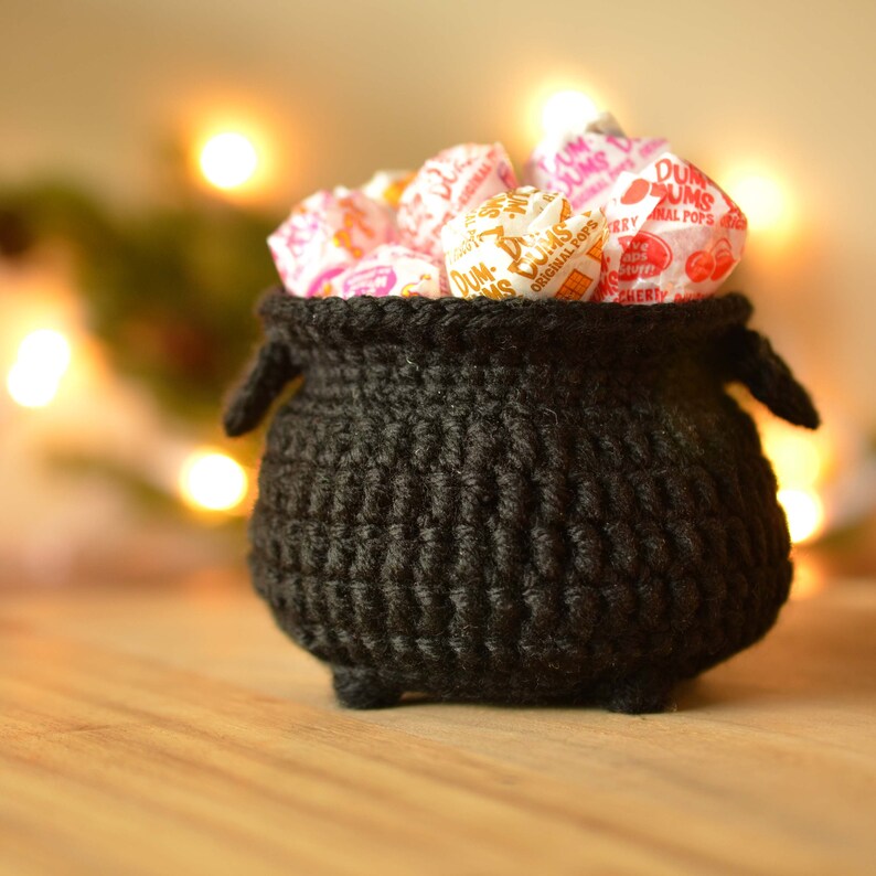 May include: A black crocheted cauldron filled with wrapped Dum-Dums lollipops. The cauldron has small handles and feet. The background is blurred with warm, out-of-focus lights, suggesting a festive or seasonal theme.