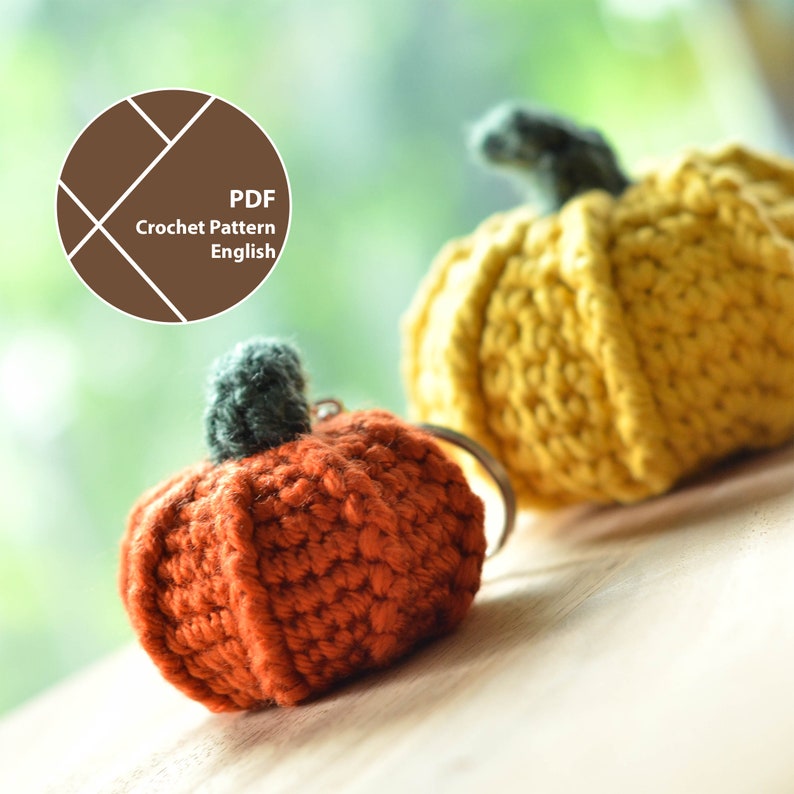 Op de afbeelding: Close-up van twee gehaakte pompoenen. E&eacute;n is oranje met een groene steel en een sleutelhanger. De andere is geel met een groene steel. De afbeelding bevat ook een bruine cirkel met de tekst "PDF Crochet Pattern English".
