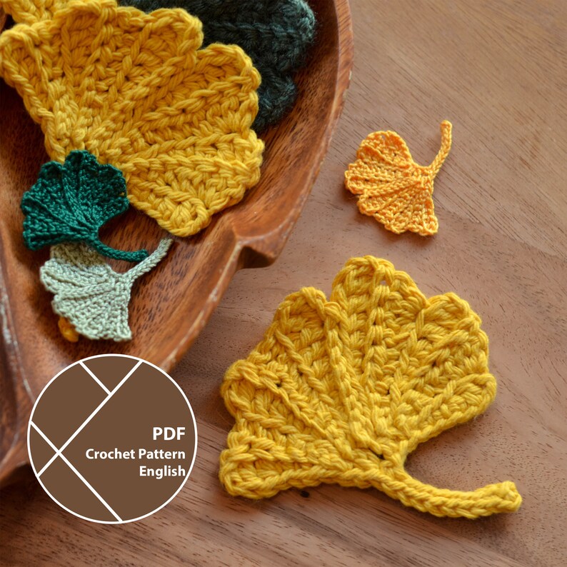 Op de afbeelding: Gehaakte ginkgo bladpatronen in tinten geel, groen en olijfgroen. De bladeren zijn gerangschikt in een houten kom op een houten oppervlak. De afbeelding bevat een afbeelding met de tekst "PDF Crochet Pattern English".