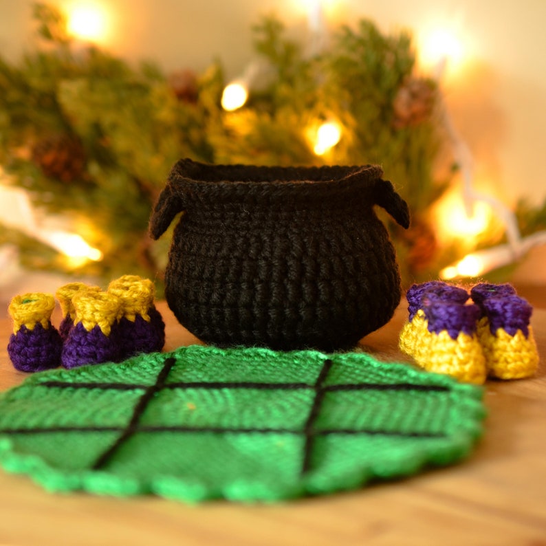 May include: A black crochet cauldron with two small handles sits on a green leaf-shaped mat. Three pairs of purple and gold booties are arranged around the cauldron. The background features a blurred display of lights and greenery.