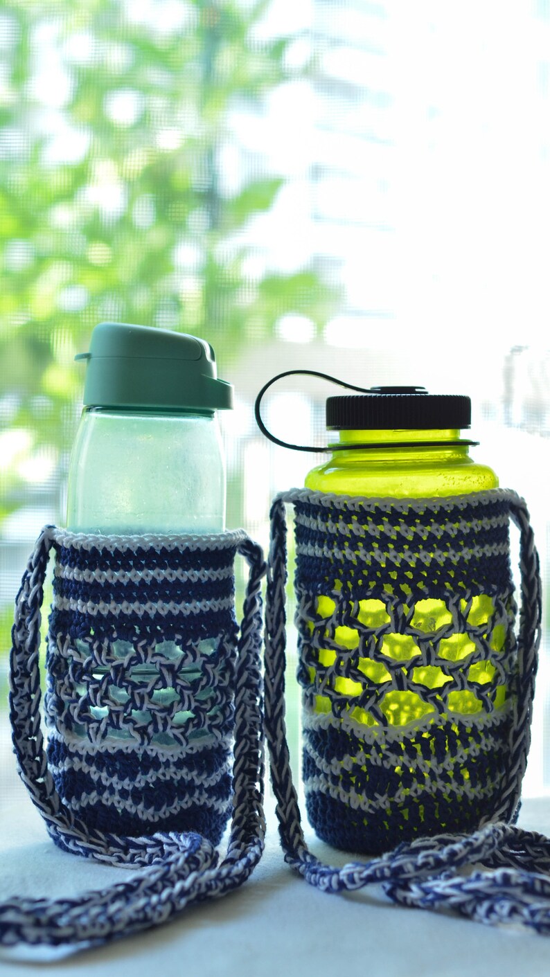May include: Two water bottles with crochet bottle holders. One bottle has a green lid, the other a black lid. The holders are blue and white, with a braided strap. The bottles are on a white surface, with a blurred green background.