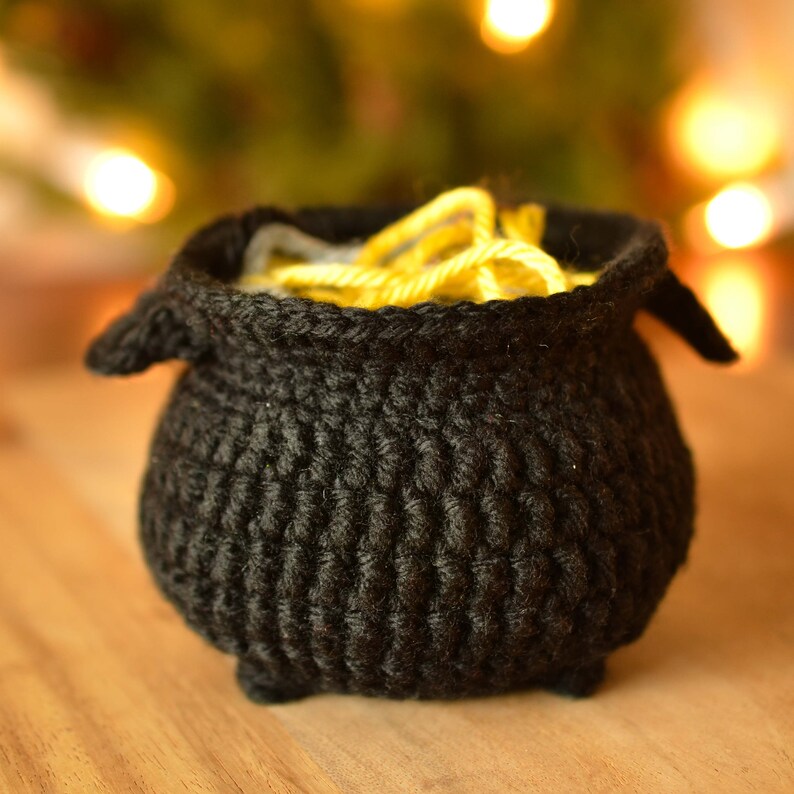 May include: A handmade black crochet cauldron with yellow yarn inside. The cauldron has a rounded body, small feet, and two handles. The yarn adds a pop of color against the dark background. This item is perfect for Halloween or fantasy-themed decor.