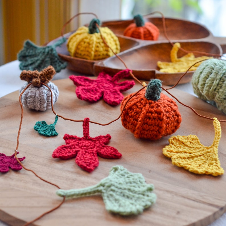 Op de afbeelding: Een handgemaakte herfstslingers met gehaakte pompoenen en bladeren in verschillende kleuren. De slinger bevat oranje, gele, rode, groene en grijze elementen, geregen met een bruin koord. De pompoenen hebben groene stelen.